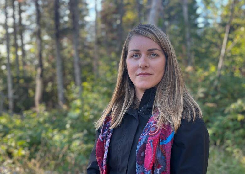 A woman in a coat and scarf stands with autumn trees in the background.