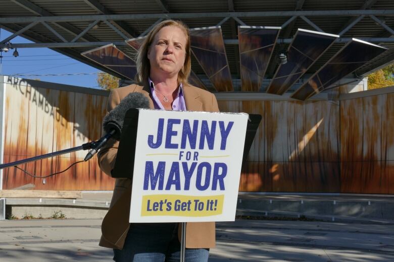 A woman wearing a suit is standing a podium and speaking into a microphone, with a pavilion in the background.