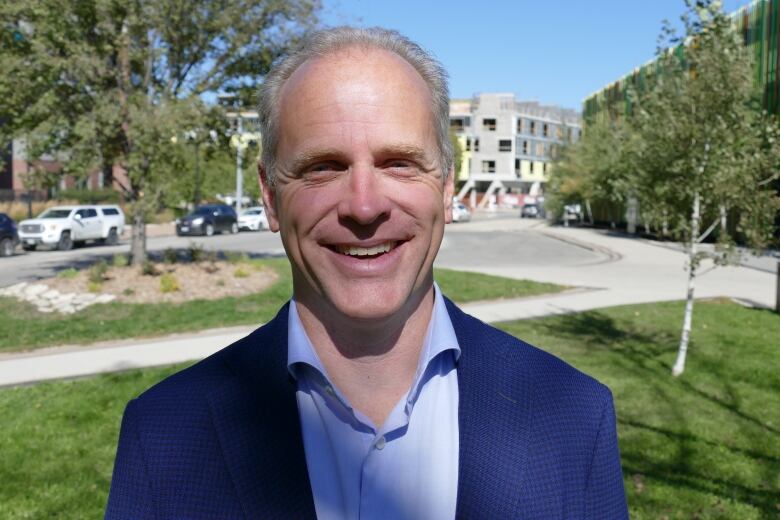 A smiling man wearing a blue suit jacket and shirt is standing in a park with a construction site in the background.