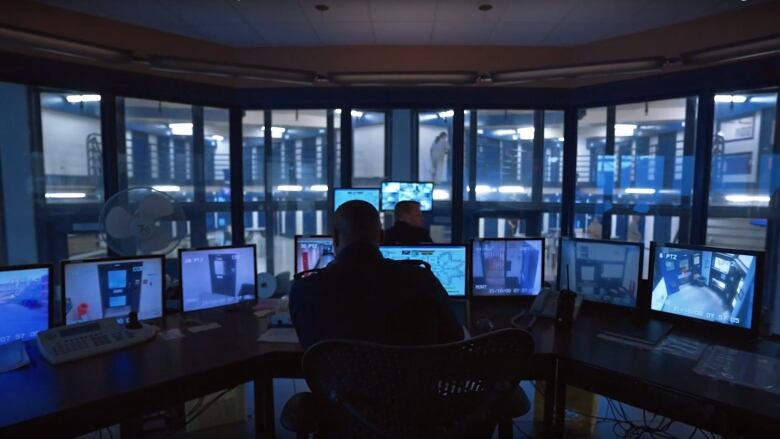 The back of a man in uniform can be seen as he sits in a glassed-in area of a larger building, looking at multiple screens with images of various areas of a corrections facility.