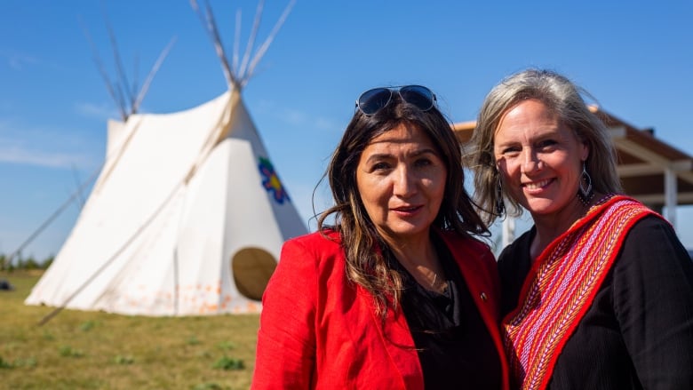 Two women stand together smiling in front of teepee.