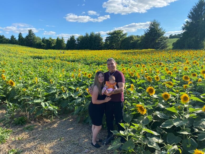 Two smiling people stand in a sunflower patch, holding a baby. 