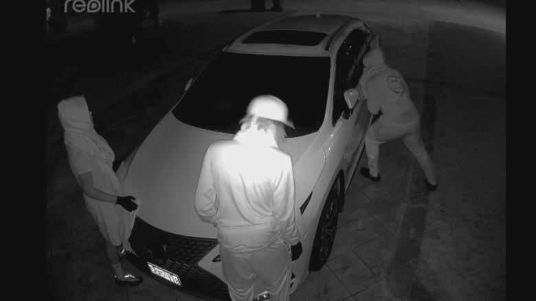 A screenshot from video shows three people wearing hooded clothing and gloves attempting to steal a car from a driveway. 