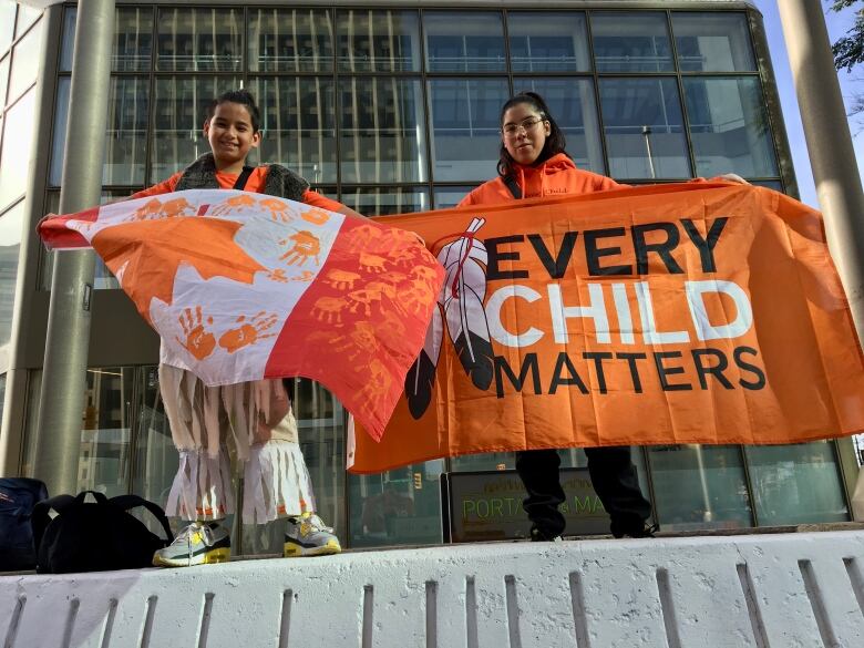 Two youth hold up flags - one is an upside down Canada flag with orange hand prints, the other says 