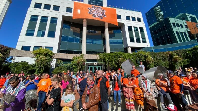 A crowd of people outside a large building with and Every Child Matters flag hanging from it