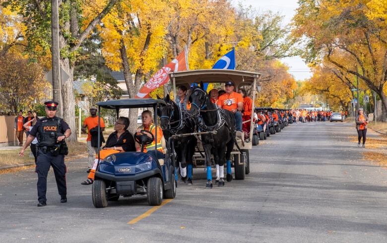 A police officer escorts the leaders of a long parade of people, many of whom are wearing orange, driving golf carts and walking outside. 