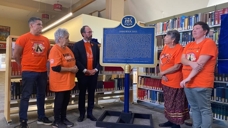 Five people wearing orange shirts stand around a new heritage plaque inside a library.