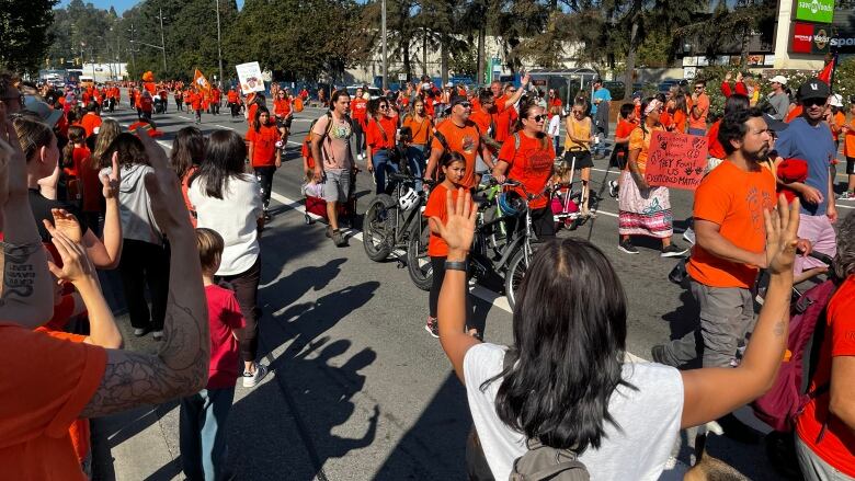 People lined up along a sidewalk hold up their hands as hundreds of people walk past wearing orange garments.