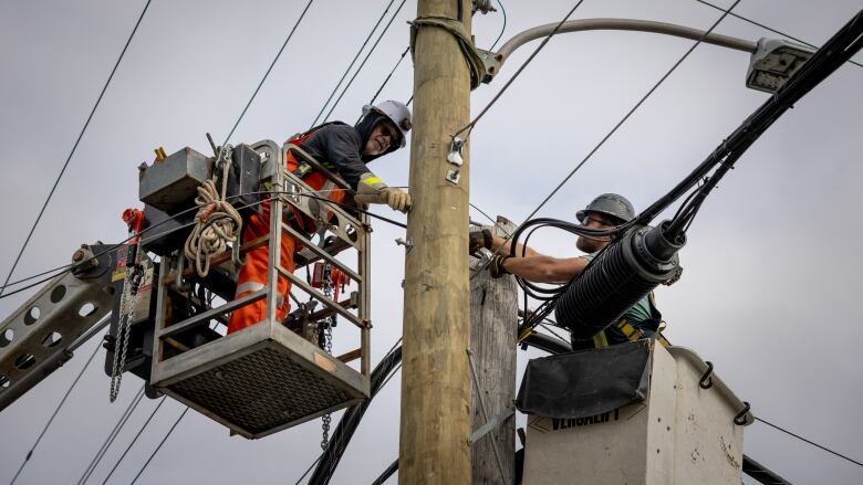 Two men wearing hard hats are seen on either side of a power pole. Both are in box lifts and have their arms outstretched toward the pole wile they hold tools.