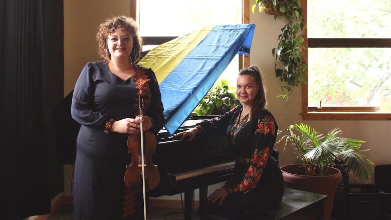 A woman holding a violin stands next to a grand piano, with another woman seated on the bench. A Ukrainian flag is draped on the open lid of the piano.