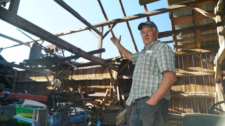 David Spence hold up his right arm, pointing to the missing roofing on his machine shed. Tractors and other farm equipment are visible inside the shed.