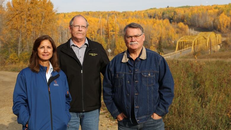 Three people stand on a bank with an old yellow bridge behind them.