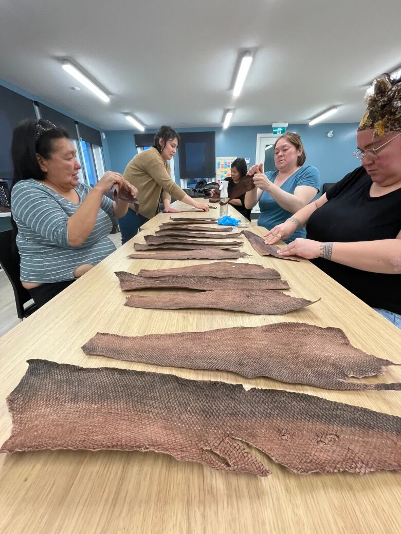 Women stand and sit on both sides of a long table lined with dark brown fish skins.