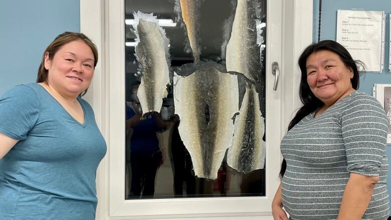 Two women smile at the camera on either side of a window, which features five flat fish skins stuck to the glass.