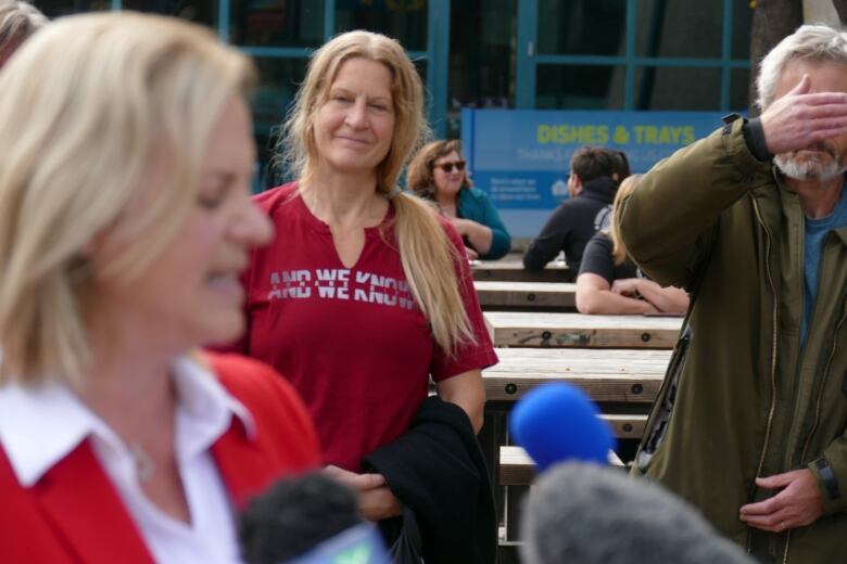 A woman on the left is speaking into microphones, while behind her another woman is smiling and looking at her. Beside her, a man is standing holding his hand in front of his face.