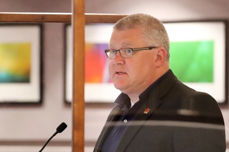 A man with short hair wearing glasses speaking at a microphone. 