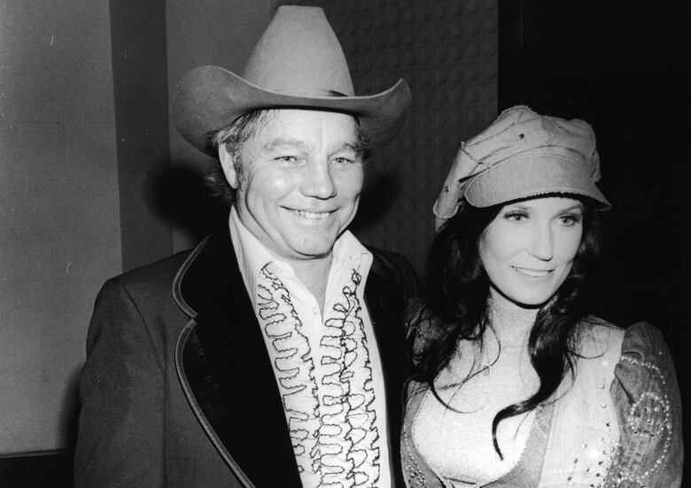 A man and woman pose together in a black and white archival photo. The man has his arm around the woman. The man wears a tall cowboy hat, frilled white shirt and a jacket with wide lapels. The woman on his right wears a large pageboy hat, and bejeweled denim jacket and pants. 