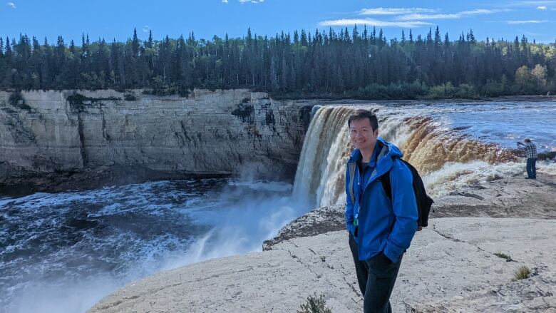 Smiling man in blue coat in front of massive waterfalls, rocks, forest.