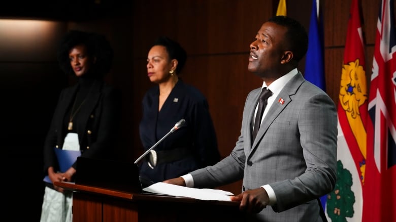 A man stands in front of podium talking. Two people stand behind him. 