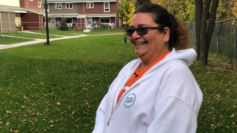 A woman wearing a white sweater is standing on a lawn with trees and a row of townhouses in the background.