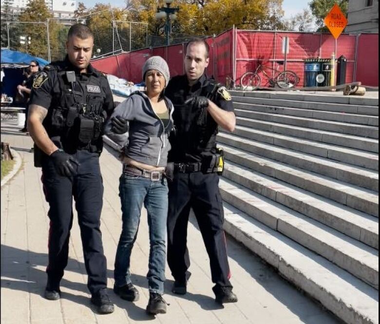 Two male Winnipeg police officers flank a woman in jeans, wearing a grey tuque, whose hands are handcuffed behind her back.