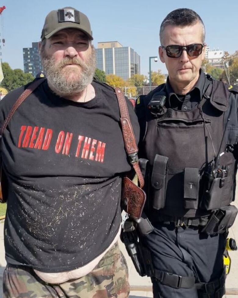 A Winnipeg police officer holds the left arm of a bearded man with handcuffs behind his back, wearing camouflage pants, a black shirt with the words TREAD ON THEM printed in red, and a baseball cap bearing an upside-down Canadian flag.