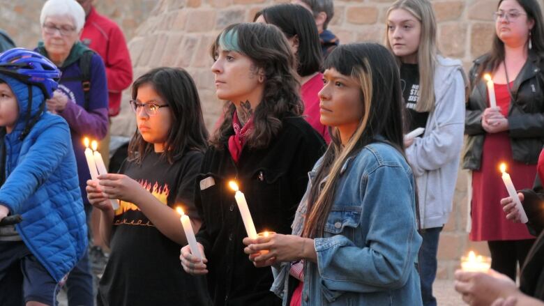 Three people with long dark hair hold candles and look solemn.