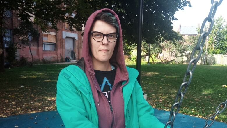 A woman with short hair and wearing a hoodie holds the chains of a park swing. She has glasses and a nose ring.