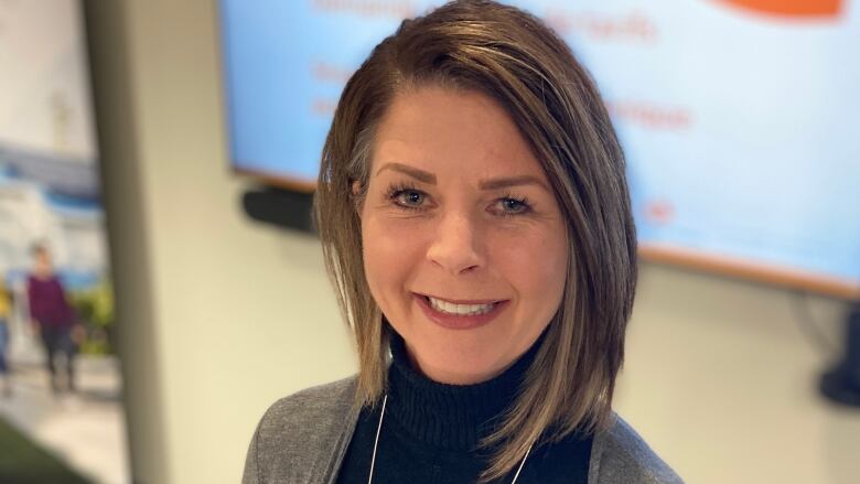 A woman wears a black turtleneck and grey sweater, and smiles directly at the camera. 