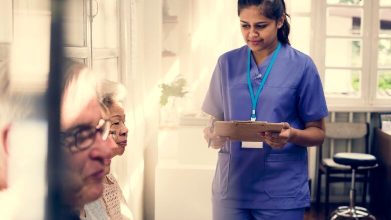 A nurse in a blue uniform stands holding a clipboard, while two elderly patients sit in a waiting room.