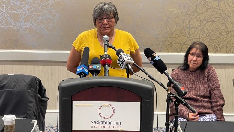 A woman in a yellow T-shirt speaks at a podium in a conference room.