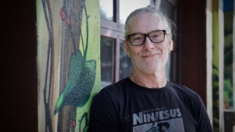 An old man with grey hair, tied back in a bun, smiles in front of a patterned pillar.