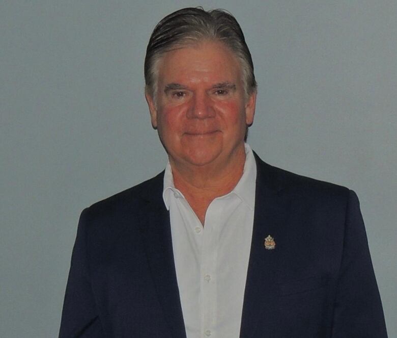 A man with short hair in a suit stands against a grey wall. 