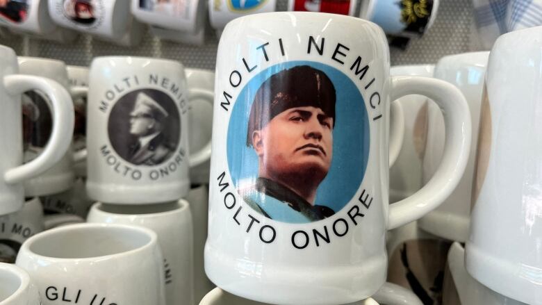 Mugs with the likeness of Benito Mussolini printed on them are stacked together on a shelf.