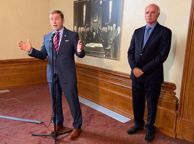 Two people, both wearing suits, stand in front of a wall. The one on the left speaks into a microphone while gesticulating. The one on the right folds his hands.