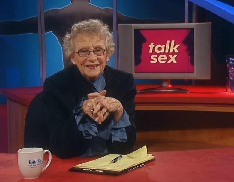 An elderly woman sits at a desk with a pen and pad of paper. A screen behind her showing the words 