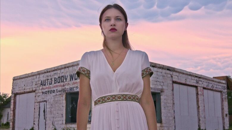 A woman in a white dress with a Ukrainian-print belt stands outside a building with peeling white paint and a partially visible sign reading 