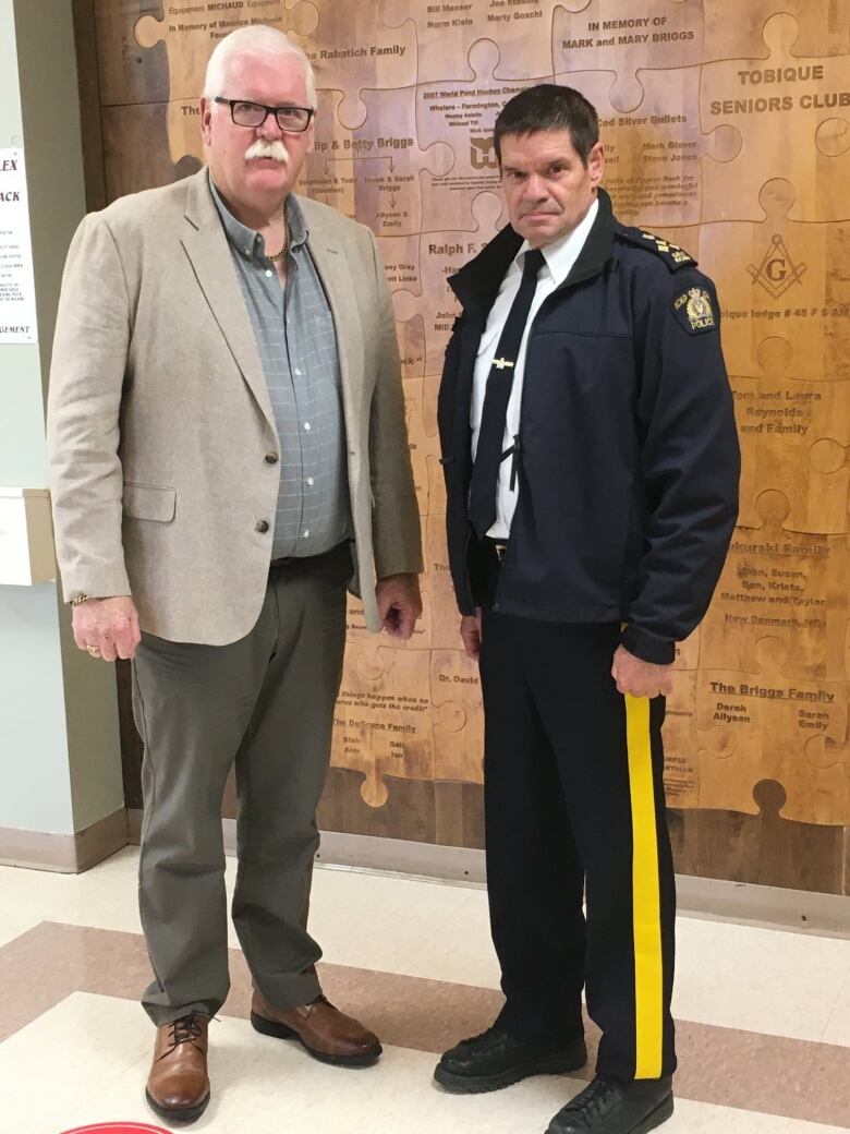 Two individuals, standing side by side, staring seriously. One dressed in a suit, the other dressed in an RCMP uniform.