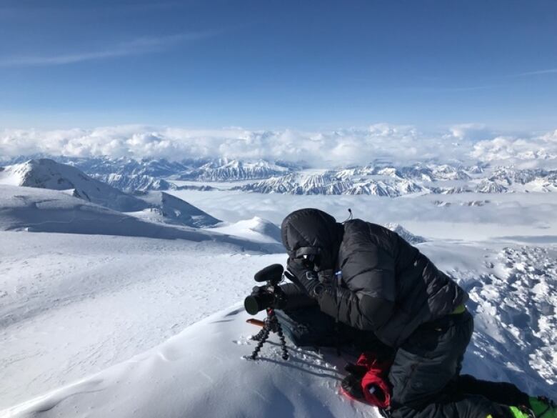 A man in winter gear with a camera and tripod, sitting atop a snowy mountain ridge.