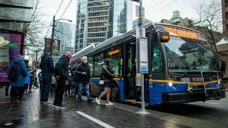 People are seen boarding a bus, wearing masks, on a rainy day.