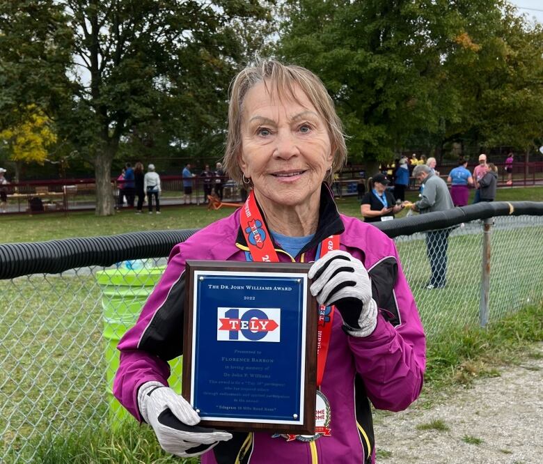 An older woman smiles into the camera. She holds up an award.