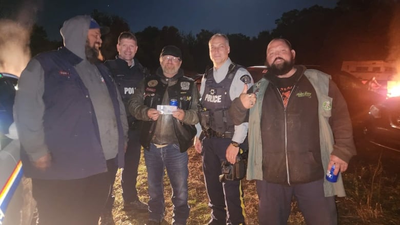 The image shows two officers wearing vests standing between three of the groups members, one of which is holding $5 bill. One of the officers has a thin blue line patch on the front of his vest. The five men are looking towards the camera and smiling.