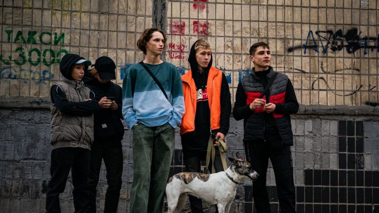 Five teenagers and a dog on a leash stand next to a graffiti-covered wall staring off in the same direction. 