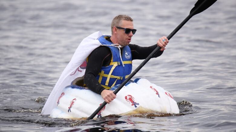 A man sits inside a giant pumpkin and paddles it through the water. 