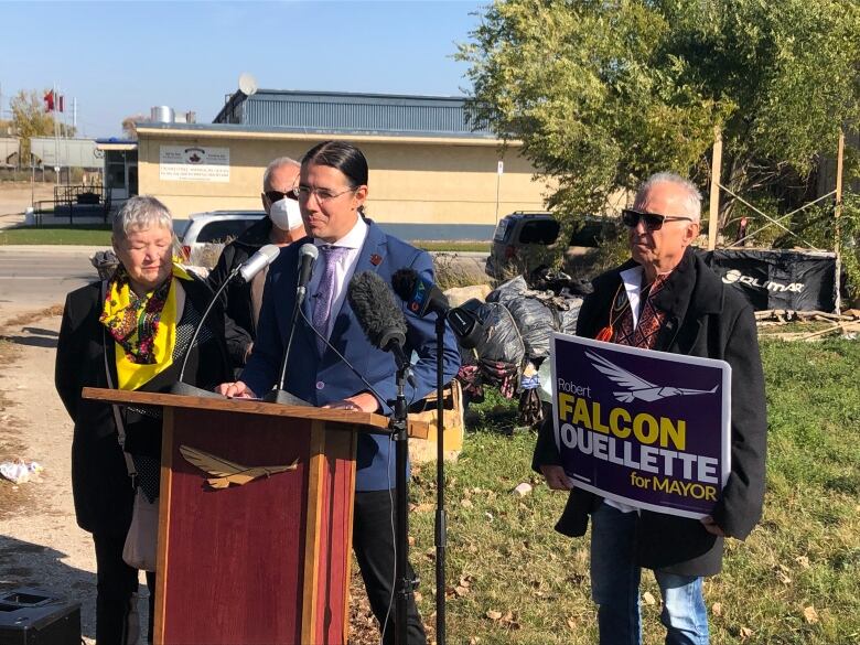 A man is speaking at a podium while three other people stand around him. Garbage and debris can be seen piled up behind them.