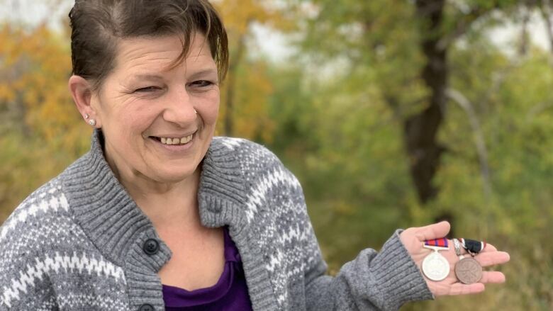 A woman in a purple shirt and a knit sweater holds a silver and bronze medal in her hand.