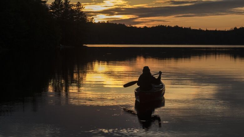 A silhouette of someone in a canoe on a lake at sunset.