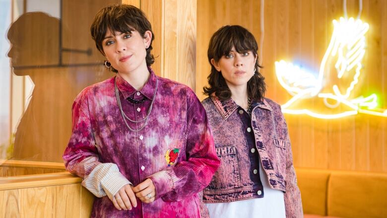 A woman with short brown hair stands next to her identical twin in a light wood-panelled room.