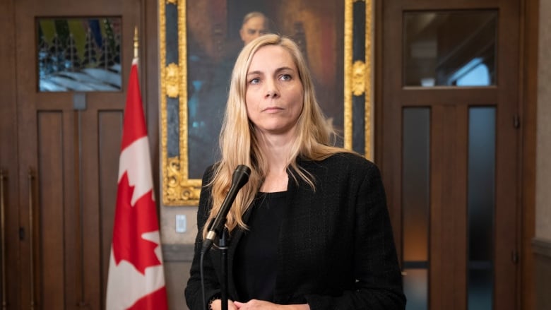 A blonde woman wearing a black top and blazer stands in front of a microphone with a Canadian flag in the background.