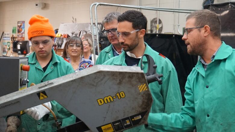 People stand behind and watch as a man works with a piece of heavy manufacturing machinery. 